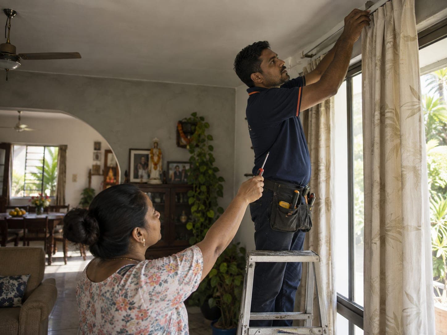 A handyman on a stepladder fixing a curtain rail while the homeowner hands up a screwdriver